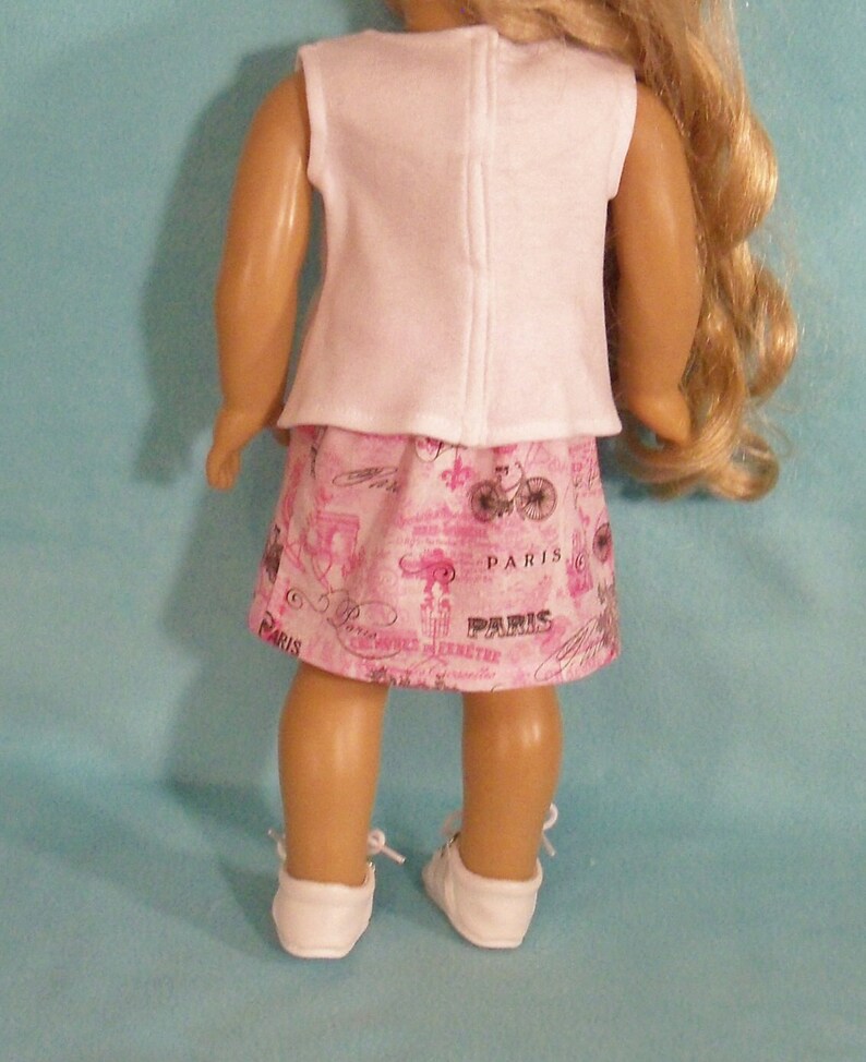 May include: A pink and white doll outfit with a white sleeveless top and a pink skirt with a Parisian print. The skirt has the word "Paris" printed on it multiple times.
