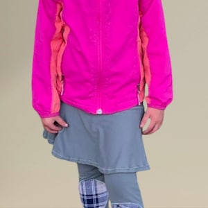 Puede incluir: Una niña lleva una chaqueta rosa con la palabra "Columbia" en la parte delantera, una falda gris y leggings grises con parches de cuadros en las rodillas.