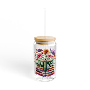 May include: A clear glass tumbler with a bamboo lid and a clear straw. The tumbler features a colorful floral design with an open book that reads "Reading is my therapy."