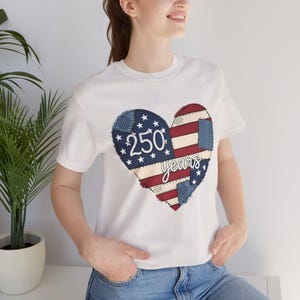 May include: A white short-sleeved t-shirt featuring a large heart-shaped graphic on the chest. The heart design mimics a patchwork quilt pattern with red and white stripes and a blue section with white stars. The text 250 years is printed in white.