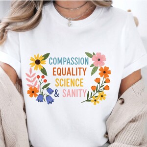 May include: White t-shirt with the words "COMPASSION EQUALITY SCIENCE & SANITY" in colorful text, surrounded by a floral border of yellow, orange, pink, and blue flowers and green leaves.