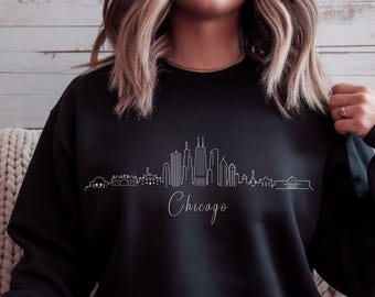 May include: Black sweatshirt featuring a white line drawing of the Chicago skyline. The word "Chicago" is written in cursive below the skyline. The sweatshirt is a solid black color.