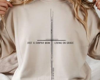 May include: Beige sweatshirt with a black cross design. The text reads "JUST A SUBPAR MOM LIVING ON GRACE" with "JOHN 19:26-27" printed vertically. The sweatshirt is a neutral color and has a crew neck.
