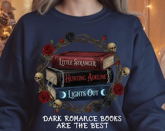 May include: Navy blue sweatshirt featuring a stack of three books with the titles "Little Stranger", "Hunting Adeline", and "Lights Out". The books are surrounded by a wreath of roses, skulls, and vines. The text "Dark Romance Books Are The Best" is below the books.