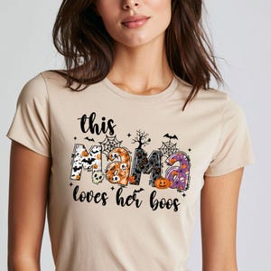 May include: A beige short-sleeved t-shirt featuring a graphic print with the text this MAMA loves her boos. The letters are filled with Halloween patterns including pumpkins, skulls, bats, spiderwebs, and ghosts in orange, black, and purple.