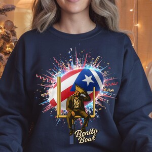 May include: Navy blue sweatshirt featuring a graphic of a man sitting on a goalpost with the Puerto Rican flag as the football. The text "Benito Bowl" is below the graphic. Confetti and celebratory elements surround the design.