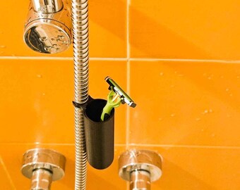 May include: A black shower razor holder attached to a chrome shower hose. A green and black razor is stored inside the holder. The background is an orange tiled wall and chrome shower fixtures.