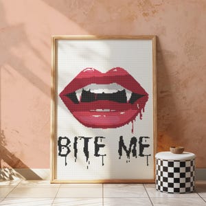 Puede incluir: Un diseño de punto de cruz enmarcado en blanco con una boca roja abierta con colmillos y el texto "Bite Me".