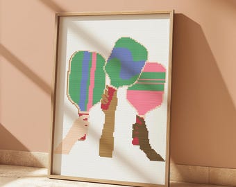 May include: A framed cross-stitch artwork featuring three colorful paddleball rackets held by three hands. The rackets are green, blue, and pink with white stripes. The hands are brown, tan, and white.