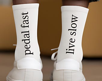 May include: A pair of white socks with the words "pedal fast" and "live slow" printed in black. The socks are worn with white sneakers. The image is a close-up of the feet and lower legs.