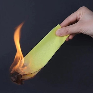 May include: A hand holding a yellow strip of material, with flames consuming the bottom edge. The fire is bright orange and yellow against a dark background, creating a contrast between the burning material and the hand.