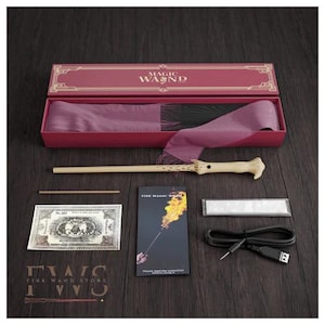May include: A magic wand set in a red box with the words "MAGIC WAND" on the lid. The set includes a wand, a prop money bill, a USB cable, and a small instruction booklet. The wand is light brown with a cream-colored handle.