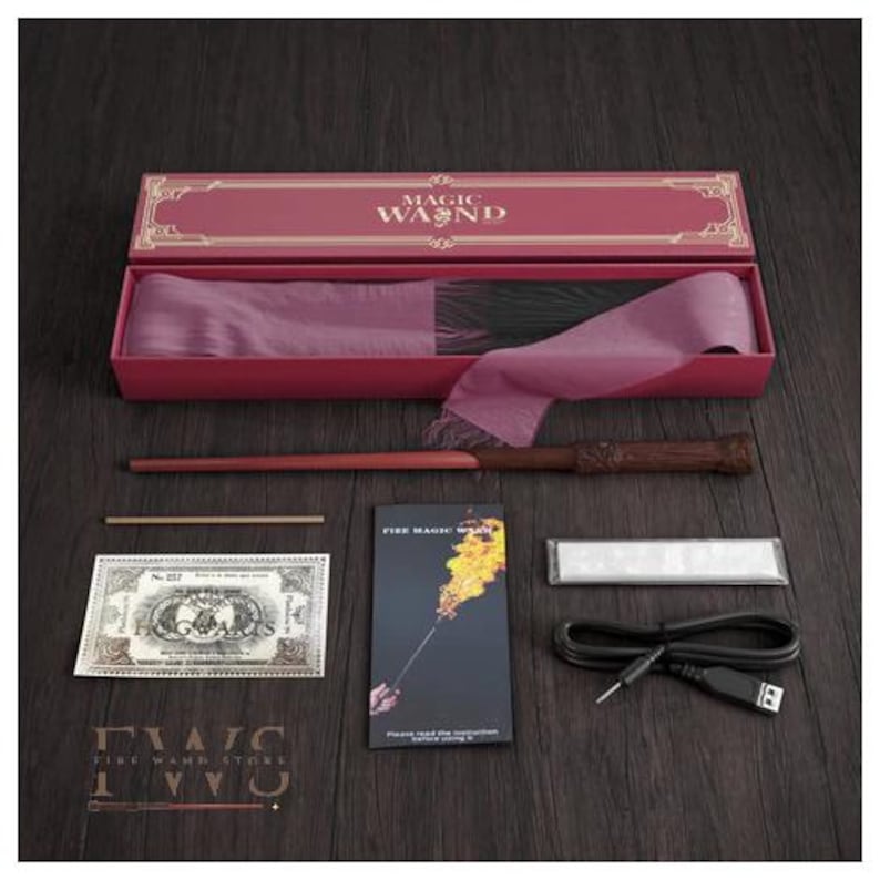May include: A magic wand set in a pink box with the words "MAGIC WAND" on the lid. The set includes a wand, a prop money bill, a small instruction booklet, a charging cable, and other accessories. The wand is brown with a detailed handle.