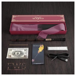 May include: A magic wand set in a pink box with the words "MAGIC WAND" on the lid. The set includes a wand, a small wooden stick, a prop money bill, a "Fire Magic Wand" instruction card, a white packet, and a black USB cable.