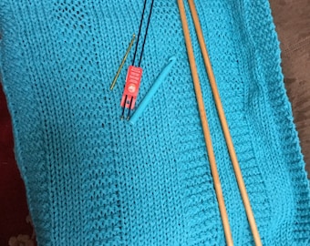 May include: A turquoise knitted blanket with wooden knitting needles and a crochet hook lying on top.