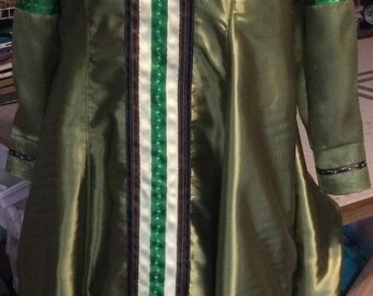 May include: Olive green satin dress with long sleeves. The dress features a central panel of white, green, and brown vertical stripes. Green ribbon accents the sleeves. Black trim outlines the hem and the central panel.