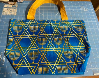 May include: A blue tote bag with a gold Star of David pattern and menorah designs. The bag has yellow handles.