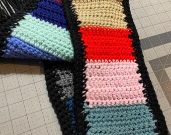 May include: A handmade crochet scarf with a colorful, patchwork design. The scarf features blocks of yellow, brown, beige, red, pink, teal, and orange, bordered with black. The scarf has black fringe.