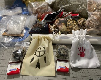 May include: A collection of small pouches and bags, some with Native American designs, filled with herbs and other items. The pouches are arranged on a white surface with a white ribbon.