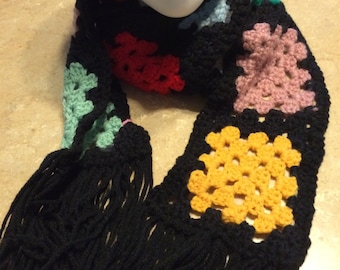 May include: A handmade crochet scarf featuring a series of colorful granny squares in shades of yellow, pink, red, and light green, all bordered by black yarn. The scarf is finished with long black fringe tassels at both ends of the garment.