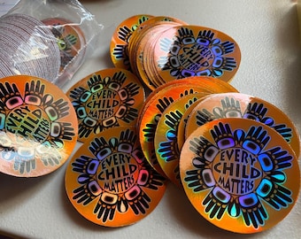 May include: A pile of iridescent gold stickers with the phrase "Every Child Matters" in a rainbow font. The stickers feature a stylized handprint design.