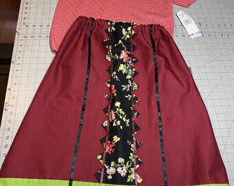 May include: A coral-colored short-sleeved sweater and a burgundy skirt with a black floral panel and green trim. The skirt has black ribbon accents and a green and gold border. The outfit is displayed on a cutting mat.
