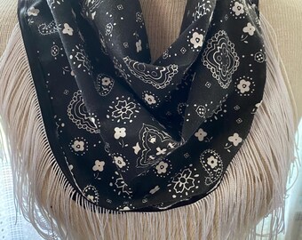 May include: A black and white patterned scarf with a white fringe trim. The scarf features a paisley and floral design. The fringe is made of long, thin white threads. The scarf is displayed on a mannequin.