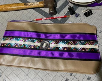 May include: A tan leather clutch with a purple, black, and white ribbon design. The ribbon has a geometric pattern and a silver button in the center.