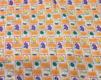 May include: Orange fabric with a repeating pattern of yellow lemons, purple grapes, and green leaves. The pattern is set within a grid of red squares.