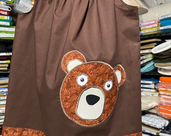 May include: A brown sleeveless dress with a brown bear applique on the front. The dress has a brown and orange floral print on the bottom hem.