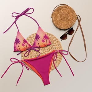 Puede incluir: Un bikini rosa y morado con estampado de palmeras. La parte superior del bikini es de estilo triangular con tirantes ajustables. La parte inferior del bikini es de estilo de amarre lateral. El bikini está sobre una estera de paja tejida.