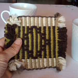 May include: A hand-woven square coaster with a geometric pattern in shades of brown, beige, and olive green. The coaster has a fringed edge and is displayed with two white ceramic mugs, suggesting a home decor item.