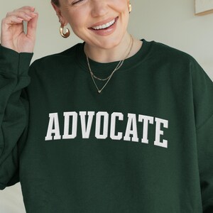 Op de afbeelding: Een donkergroene sweatshirt met het woord "ADVOCATE" in witte blokletters gedrukt. De sweatshirt heeft een ronde hals en lange mouwen. De afbeelding toont een persoon die de sweatshirt draagt.