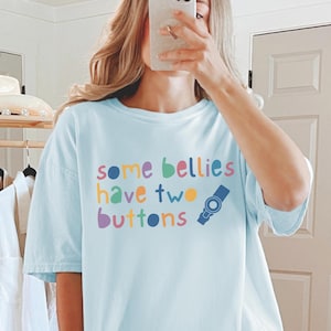 May include: A light blue t-shirt with the text "some bellies have two buttons" in rainbow colours. The text is stylised with a cartoon-like font. A small blue and white illustration of a button is next to the text.