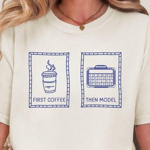 May include: Cream-colored t-shirt with a blue graphic. The graphic features a coffee cup and the text "FIRST COFFEE" on the left, and a keyboard and the text "THEN MODEL" on the right.