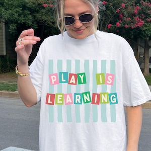May include: A white short-sleeved t-shirt featuring a graphic print of light blue vertical stripes. Over the stripes are colourful rectangular blocks containing the text PLAY IS LEARNING in pink, green, and red letters on a white background.