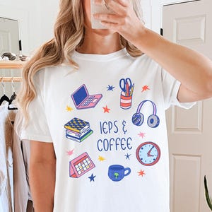 May include: White t-shirt with a colourful graphic design featuring a laptop, books, scissors, headphones, a clock, a calendar, and a coffee cup. The text "IEPs & COFFEE" is also printed on the shirt.