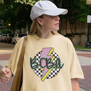 May include: A cream-coloured short-sleeve t-shirt featuring a graphic design of a black and white checkered circle, a pink and yellow lightning bolt, and the letters BCBA in a dark green script font, paired with a white baseball cap.