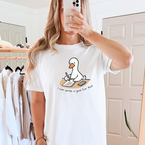 May include: White t-shirt featuring a cartoon duck writing on a notepad with a pen. The duck is sitting at a desk with a smartphone. The text on the shirt reads, "I can write a goal for that."
