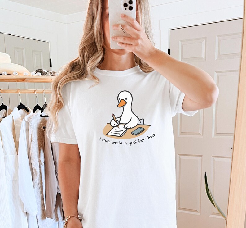 May include: White t-shirt featuring a cartoon duck writing on a notepad with a pen. The duck is sitting at a desk with a smartphone. The text on the shirt reads, "I can write a goal for that."