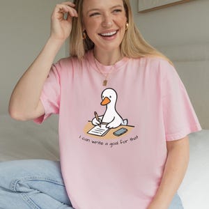 May include: A light pink t-shirt featuring a cartoon goose writing on a notepad. The goose is white with an orange beak and is sitting at a desk. The text "I can write a goal for that" is below the goose.