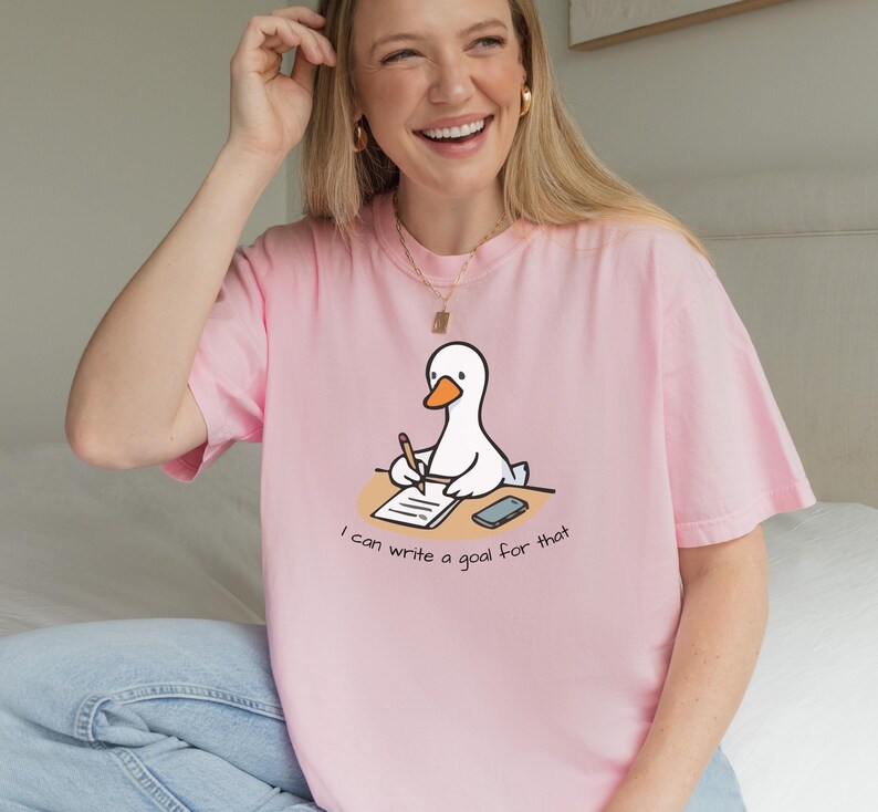 May include: A light pink t-shirt featuring a cartoon goose writing on a notepad. The goose is white with an orange beak and is sitting at a desk. The text "I can write a goal for that" is below the goose.
