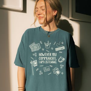 May include: Teal-colored t-shirt with the text "HOWEVER YOU COMMUNICATE, I AM LISTENING" in white, along with various white graphic icons. The icons include keyboards, a hand gesture, a computer, and playing cards. The shirt is a casual, oversized fit.