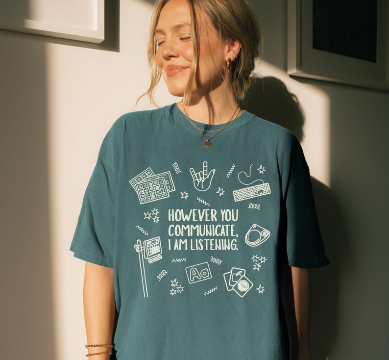 May include: Teal-colored t-shirt with the text "HOWEVER YOU COMMUNICATE, I AM LISTENING" in white, along with various white graphic icons. The icons include keyboards, a hand gesture, a computer, and playing cards. The shirt is a casual, oversized fit.