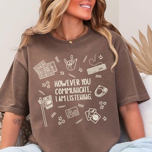 May include: Brown t-shirt with the text "HOWEVER YOU COMMUNICATE, I AM LISTENING." The shirt features white graphic icons of a keyboard, hand gestures, and other communication symbols. The shirt is a casual, comfortable style.