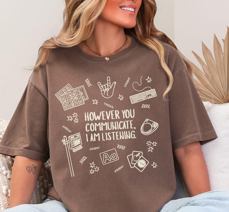 May include: Brown t-shirt with the text "HOWEVER YOU COMMUNICATE, I AM LISTENING." The shirt features white graphic icons of a keyboard, hand gestures, and other communication symbols. The shirt is a casual, comfortable style.