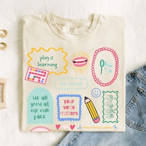 May include: Cream-colored t-shirt with colorful graphic designs. The shirt features phrases like "play is learning," "be loud," and "your voice matters." Other graphics include a board game, lips, a pencil, and hearts.