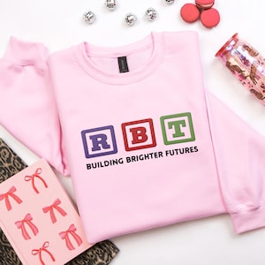 Peut inclure: Un sweat-shirt rose clair avec les lettres "RBT" dans des blocs carrés colorés, avec les mots "BUILDING BRIGHTER FUTURES" en dessous. Le sweat-shirt est entouré d'objets décoratifs, dont un gobelet, des macarons et un carnet.
