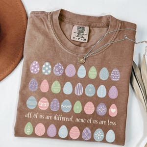 May include: A brown t-shirt with a crew neck and the text "all of us are different, none of us are less". The shirt features a graphic of colorful Easter eggs. A silver necklace with a small pendant is draped across the shirt.