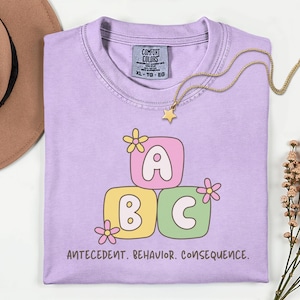 May include: A light purple Comfort Colors t-shirt with the letters "ABC" in a stacked block design, each block in a different color. The shirt also features the text "ANTECEDENT. BEHAVIOR. CONSEQUENCE." and a gold necklace with a star pendant.